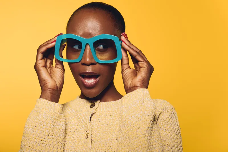 Surprised black woman with dark skin wearing oversized blue glasses and beige sweater on yellow background. Concept of fashion, expression, and vibrant portrait with confident female model.