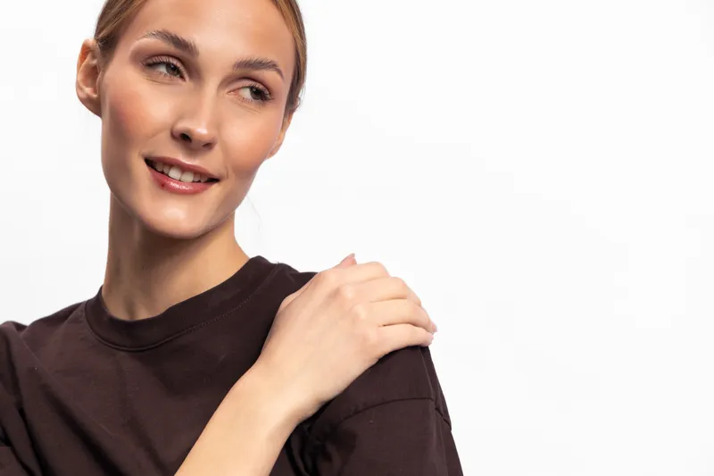 Smiling woman in brown shirt looks aside with soft expression, hand on shoulder. Studio portrait on white background. Concept for beauty, confidence, and natural skin.