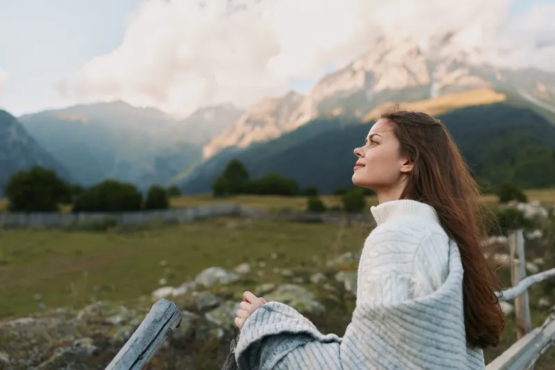 Young woman wrapped in a blanket enjoying fresh mountain air outdoors. Relaxed lifestyle concept with natural light and scenic background of hills and clouds.