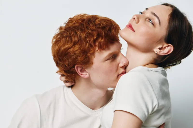 Couple, young man and woman in white shirts, close embrace on plain background. Intimate moment with tender expression and warm connection between two people with red hair.