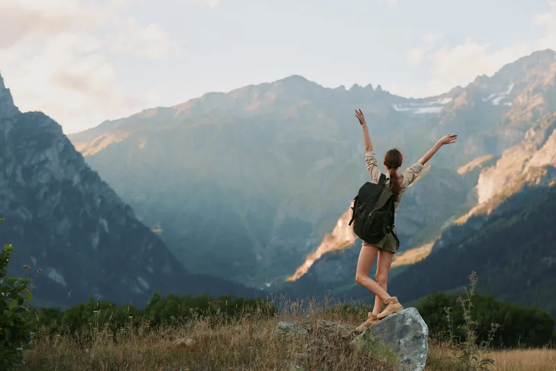 Young woman lifestyle traveler stands on rock with raised arms enjoying mountain landscape during sunny day in nature outdoors adventure concept.