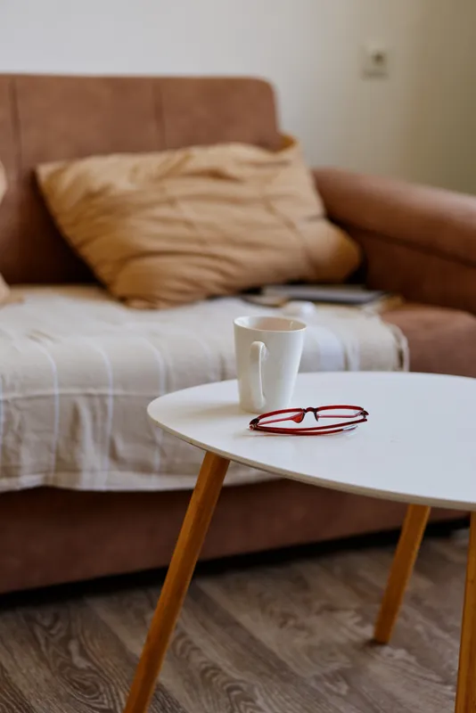Cozy living room scene with a white mug on a round table and a pair of red glasses, soft cushions on a sofa, warm natural light filtering through the room creating a calm domestic atmosphere.