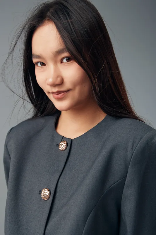 Young woman with long dark hair wearing dark gray jacket with buttons posing against light gray background with soft smile for fashion portrait style.