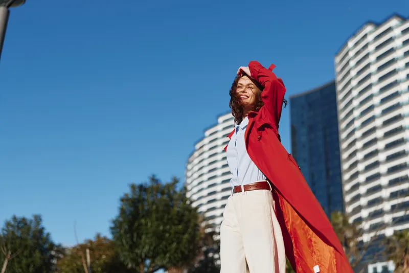 A young woman in a red cape stands on a sunny city street with arms raised in a heroic pose, skyscrapers behind and a clear blue sky, embodying confidence, freedom, and boldness.