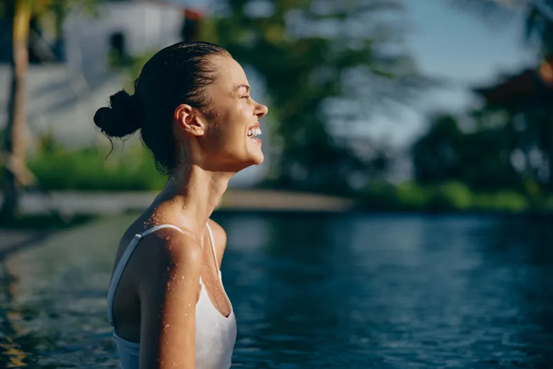 Bright joyful woman by the pool, hair in a bun, wearing a tank top, soaking in sunlight, capturing a carefree moment of summer relaxation and wellness beside refreshing water.