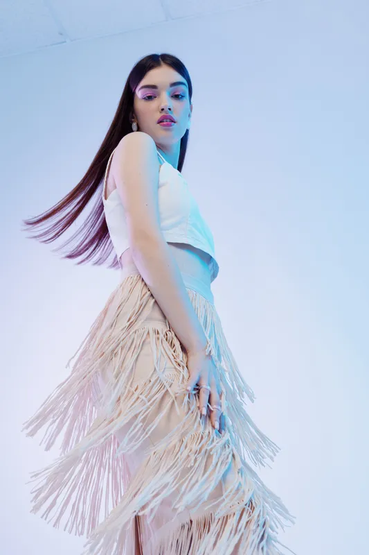 Young woman in stylish white top and beige fringe skirt poses confidently in a studio with cool blue lighting. Concept of woman beauty fashion and modern style.
