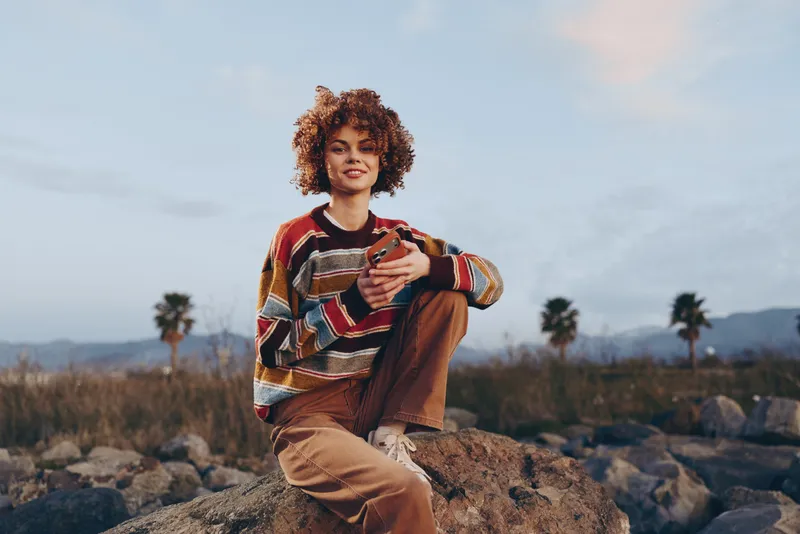 Woman lifestyle outdoors, curly hair, sweater, rocks, coat, smiling softly, sitting on stone with a relaxed pose, palms and mountains behind, nature background, soft daylight