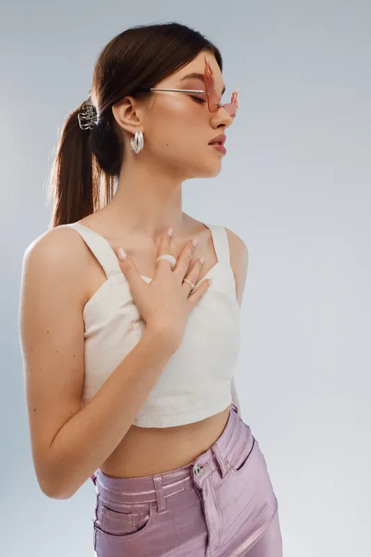 Young woman in stylish pink sunglasses and white crop top poses with hand on chest against light background. Concept of beauty, fashion, and confident lifestyle.