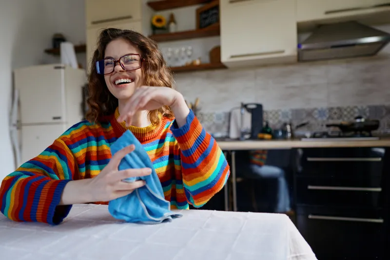 Smiling woman indoors at home, cleaning a light table with a blue cloth, vibrant striped sweater, warm kitchen background, candid lifestyle moment, natural lighting, relaxed mood