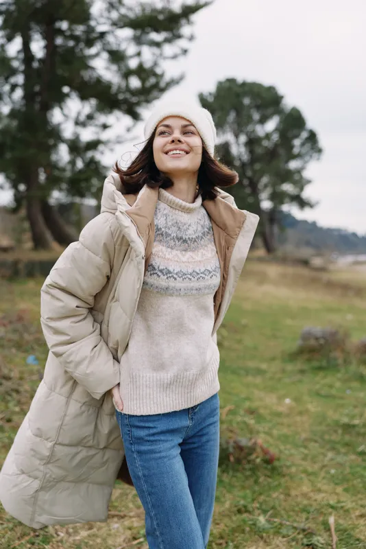 Woman in a cozy winter coat and knitted sweater outdoors, smiling in a breezy field with trees, showcasing casual seasonal fashion and carefree outdoor style.