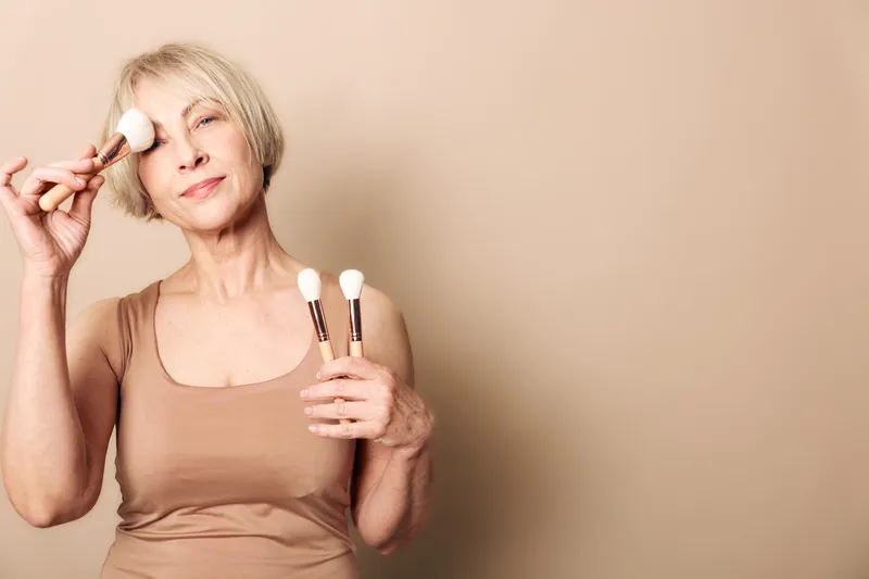 Mature woman with short blonde hair applies makeup using soft brushes on beige background. Concept of beauty routine, self-care, and natural skincare for older adults.