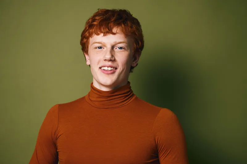 Young redhead man with curly hair wearing orange turtleneck smiling and looking at camera with friendly expression on green background. Portrait of confident male model indoors.