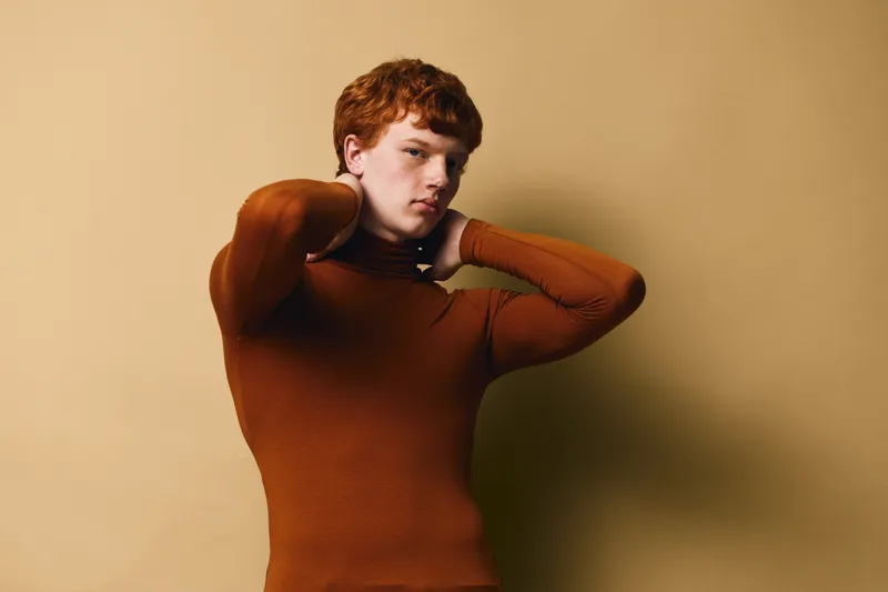 Young man with red hair wearing brown turtleneck sweater posing with hands behind neck against beige background for fashion portrait concept and styling.
