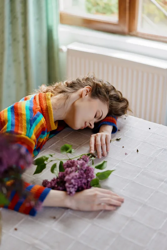 Woman resting at a sunlit table indoors, wearing a colorful sweater, flower bouquet nearby, peaceful home moment, relaxed posture, soft natural light, warm tones