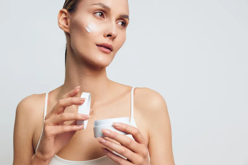 Young woman applying white face cream on cheek with fingers, holding cosmetic jar. Skincare concept, natural beauty, self-care routine, clean skin, soft lighting on neutral background.
