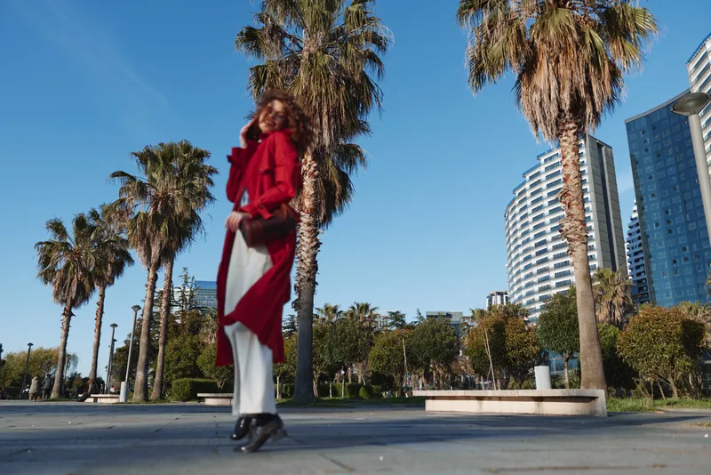 A woman in a red coat stands on a sunny urban street with palm trees and modern buildings in the background under a clear blue sky.