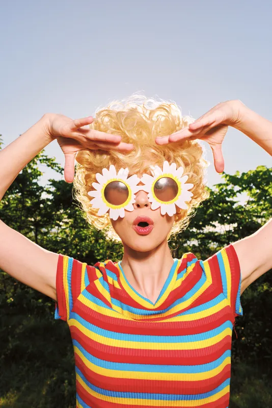 Portrait of a playful woman outdoors wearing a colorful striped top and sunflower glasses, creating a cheerful vibe. Bright sky background highlights fashion, lifestyle, and carefree energy.