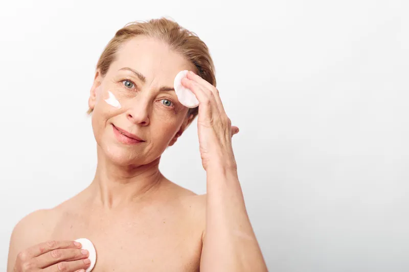 Mature woman with light skin applies skincare cream on face using cotton pads, showing natural beauty and self-care routine in bright minimal studio setting.