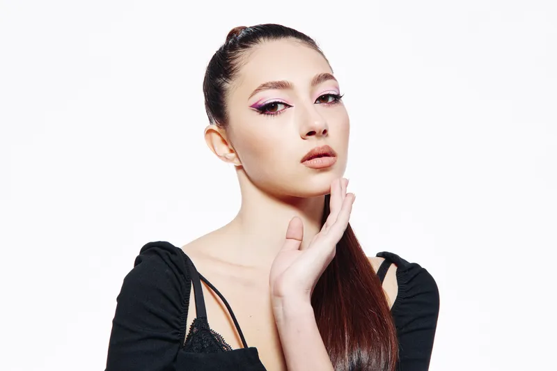 Elegant young woman with creative makeup poses wearing a black top. Shot on white background with soft lighting. Concept for fashion, beauty, minimalist portrait.