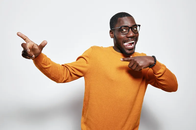 African American man with dark skin wearing glasses and orange sweater smiling and pointing to side on white background. Happy male portrait with casual style and positive expression.