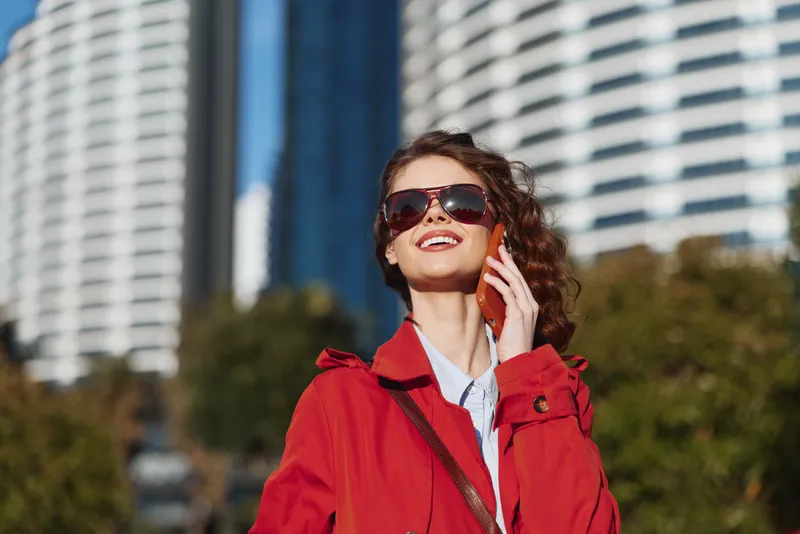 Young woman with curly hair wearing a red coat and sunglasses smiles while talking on a smartphone outdoors in an urban environment on a sunny day.