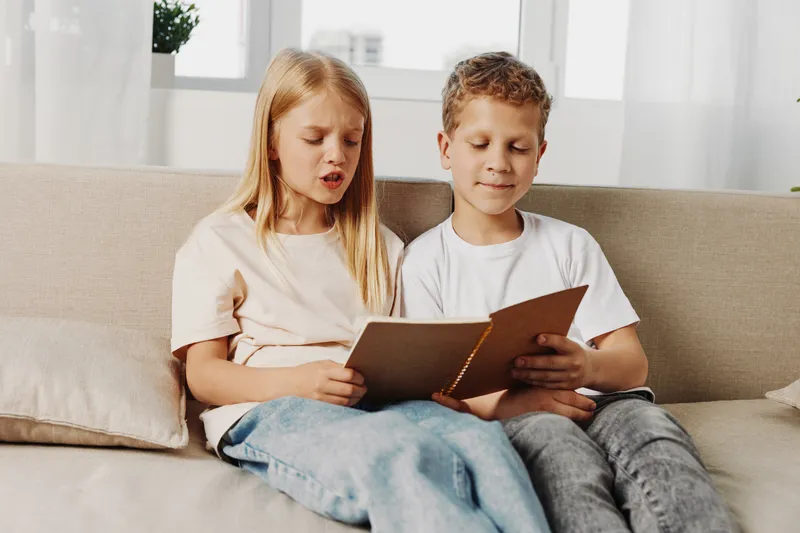 Two children sitting on a beige sofa reading a book together in a bright living room. Concept of childhood learning, education, and sibling bonding activity.