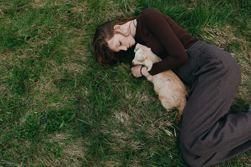 A calm lifestyle scene shows a woman lying on grass, cuddling a gentle goat kid, expressing warmth, tenderness, and connection with nature in an outdoor setting, conveying peaceful companionship and