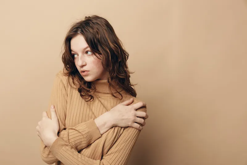 Young woman in beige sweater hugs herself and looks away with thoughtful expression. Neutral background and soft lighting create calm and introspective mood. Concept of solitude and reflection.