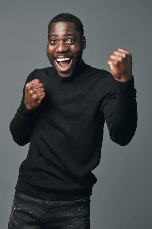 African man with dark skin wearing black sweater showing excitement and joy with clenched fists in creative conceptual visual style on gray background. Happy, energetic and expressive portrait.