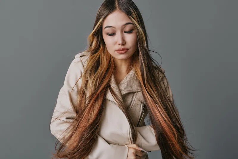 Young woman with long hair wearing casual jacket standing with eyes closed and hands in pockets against gray studio background.