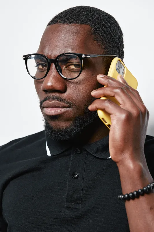 Black man with dark skin wearing glasses and black polo shirt talking on yellow smartphone with serious expression isolated on white background in studio portrait.