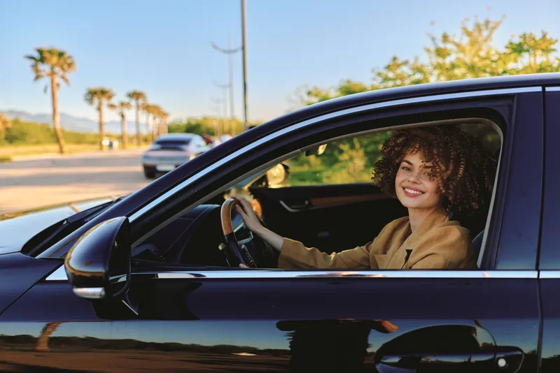 Woman driver sits in a sleek car smiling on a sunny street with palm trees, showcasing a casual lifestyle and retro film color palette, vibrant street scene. Rich vibe.
