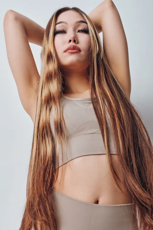 Young woman with very long straight hair wearing a sleeveless top posing with arms raised on white background for beauty and fashion portrait.