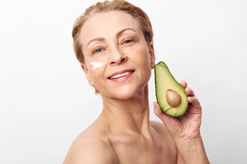 Smiling mature woman with natural skin holds fresh avocado near face with cream on cheek. Concept of healthy skincare, natural beauty, organic cosmetics, wellness, and self-care routine.