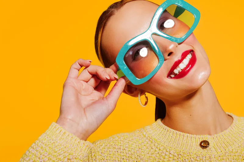 Smiling woman with large blue sunglasses and red lipstick poses against bright yellow background. She wears a yellow sweater and gold hoop earrings. Concept of fashion and summer style.