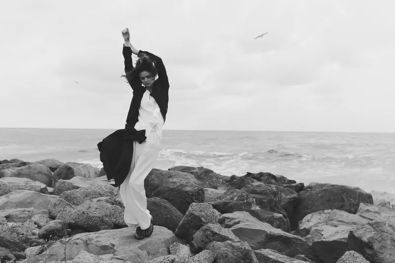 Woman leaping on rocky shore beside the ocean, white dress fluttering and black jacket streaming, dynamic motion against a windy coastal scene, dramatic balance and freedom in a monochrome setting
