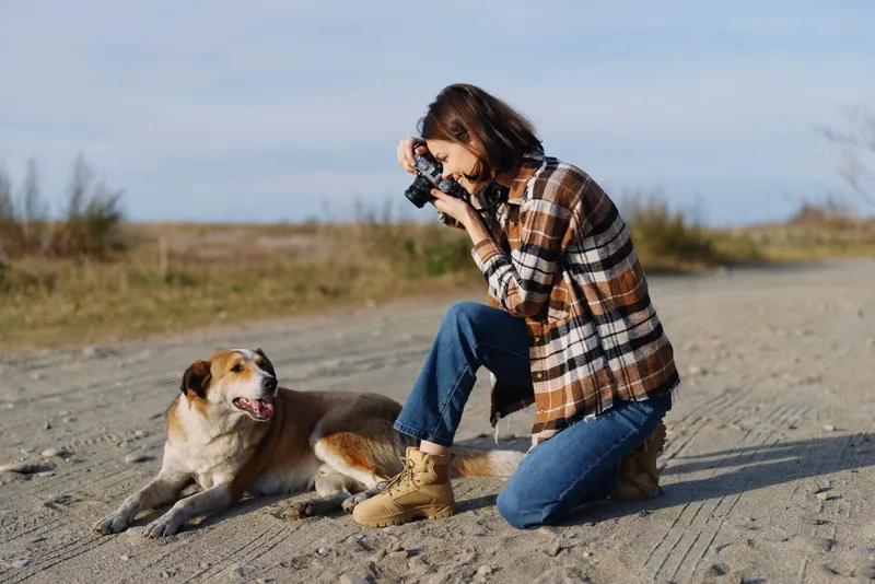 Photographer kneeling outdoors with a camera, capturing a playful dog on a sandy trail, nature surroundings, casual plaid shirt, jeans, sunlit moments and quiet companionship.