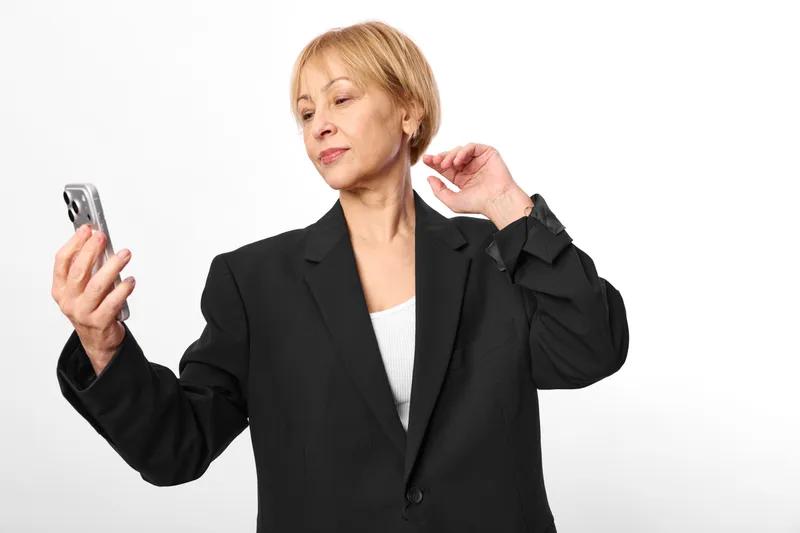 Elderly woman in a black blazer holds a smartphone and uses a hand gesture, showing communication and technology for business, studio portrait on white background.