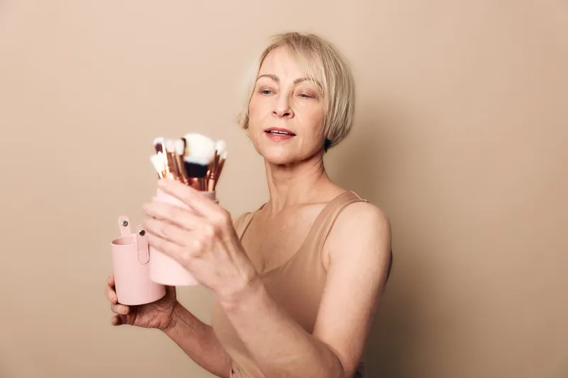 Mature woman with short blonde hair holds pink container with makeup brushes, standing against beige background. Concept of beauty routine, cosmetics tools, and skincare at home.