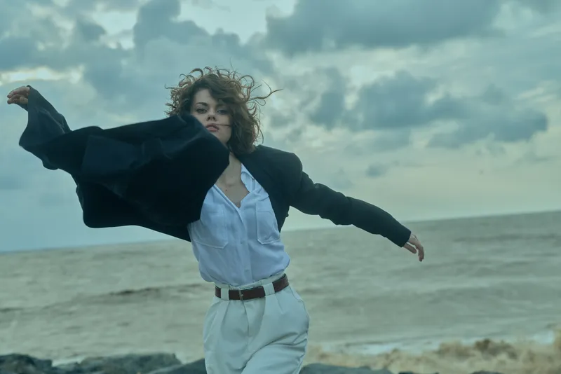 Windy beach scene with a woman in white suit and dark jacket fluttering, embracing freedom and rapid motion against a dramatic cloudy sea backdrop