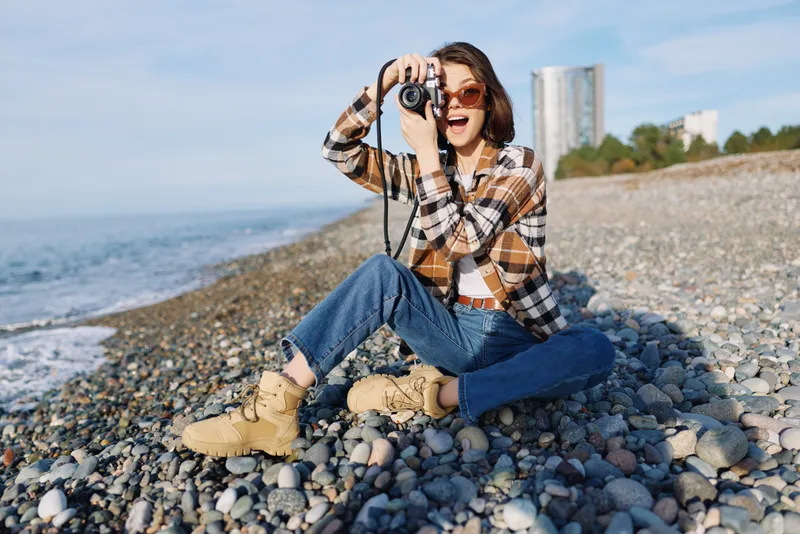 Photographer, woman on a pebble beach capturing scenes with a handheld camera, wearing plaid shirt, jeans and boots, sunlit sea in the background, candid outdoor lifestyle moment