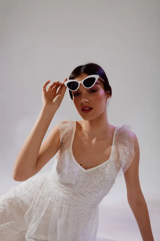 Young woman in a white sparkling dress adjusts stylish sunglasses. Her makeup is elegant and her hair is neatly styled. Concept for beauty fashion and glamour.