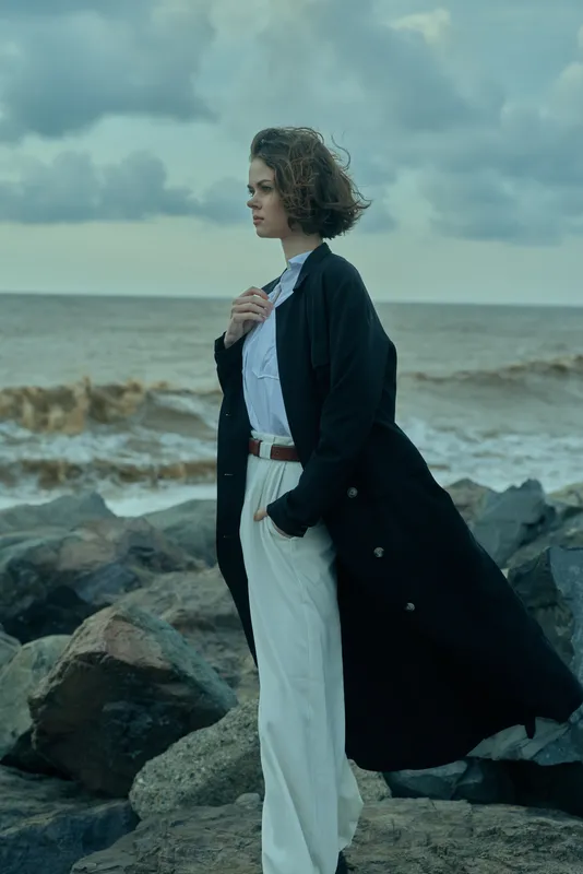 Fashionable model stands on a rocky coastline, a dark coat billowing in the wind, paired with white pants and a pale shirt, creating seaside elegance against the ocean backdrop