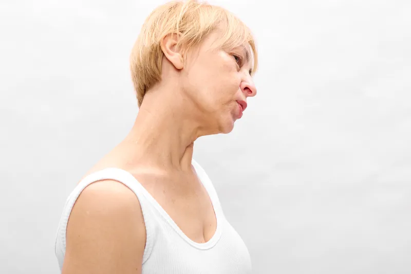 Woman portrait profile with blond hair and sleeveless white top, licking lips in a relaxed expression, soft light, plain studio background, calm mood