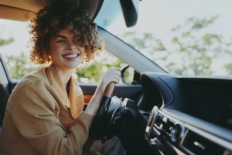 A smiling woman sits behind the wheel in a cozy jacket, daylight streams through the car window, giving retro film color tones. Concept: everyday independence, travel, and female lifestyle.