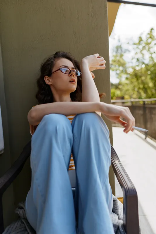 Relaxed woman with glasses sits on a balcony, leaning against a pillar, wearing blue pants, looking away. Casual lifestyle moment with natural daylight and a serene mood. Soft lighting emphasizes