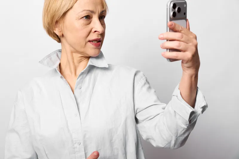 Elderly woman holds smartphone for a selfie, side profile on a white background, focused expression, home portrait, technology social communication concept