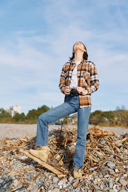 Outdoor fashion portrait of a woman standing on a rocky beach, head tilted upward, wearing a plaid shirt, jeans and boots, embracing open sky and natural light