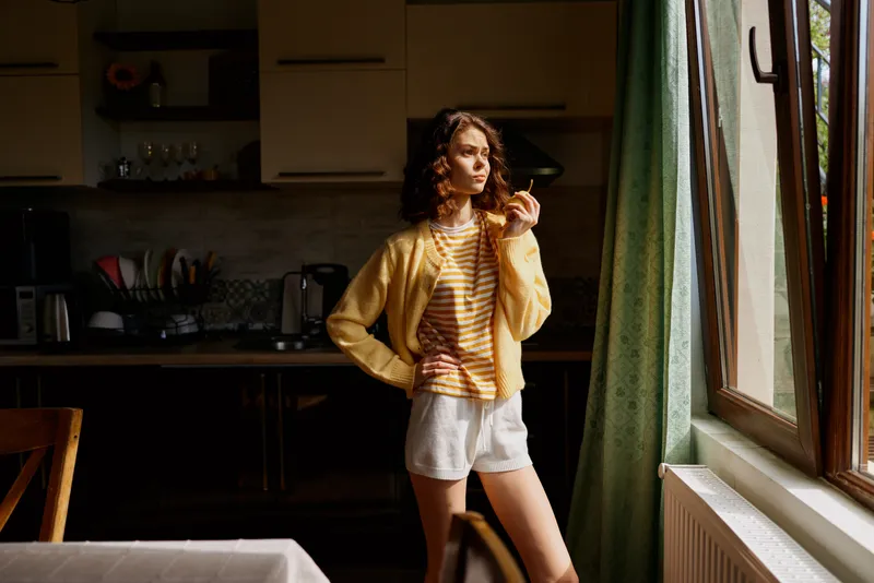 A young woman stands in a sunlit kitchen by a window, wearing a yellow cardigan over a striped shirt and white shorts, posing confidently with one hand on her hip.
