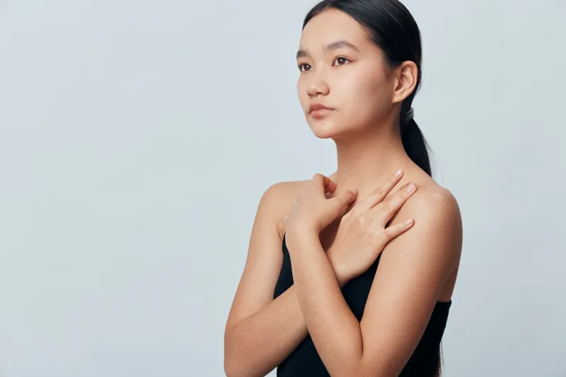 Young woman with clear skin and black hair tied back wearing a black top with crossed hands on chest posing against plain light background in studio portrait with calm expression.